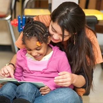 A little girl sitting on a woman's lap reading a book