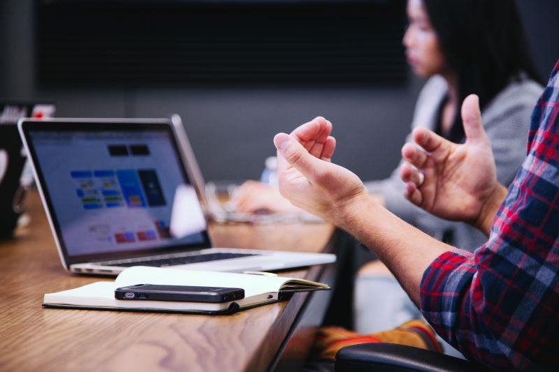 Person using hand gestures during a conversation while working on a laptop with another person in the background.