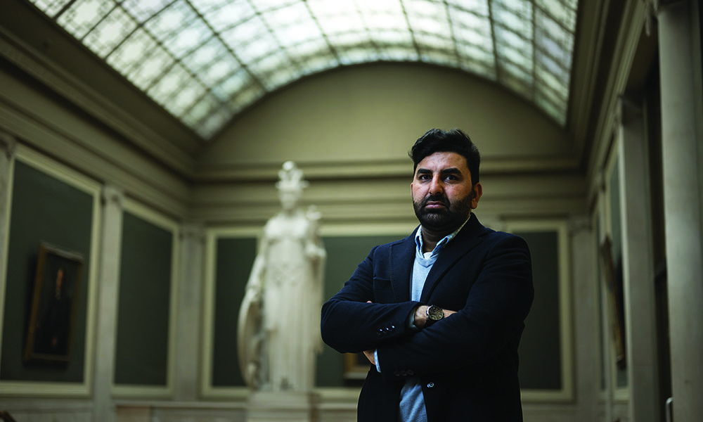 Hameedi standing with arms folded in front of statue in Rush Rhees Library.